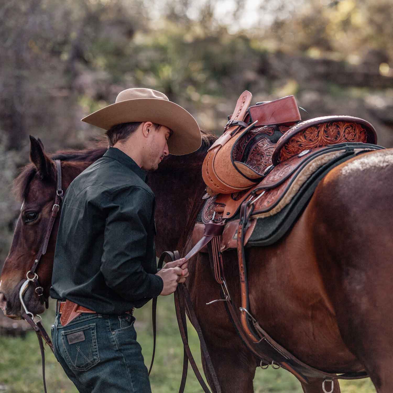 A young cowboy saddling his horse with a western saddle on in the forest